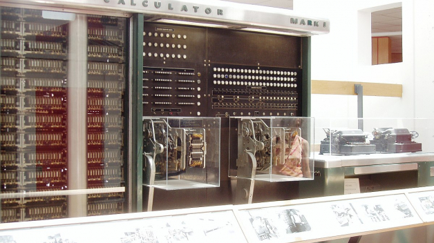 Mark I. nepřipomíná dnešní počítače ani vzdáleně. V pozadí jsou vidět výstupní zařízení – psací stroje.